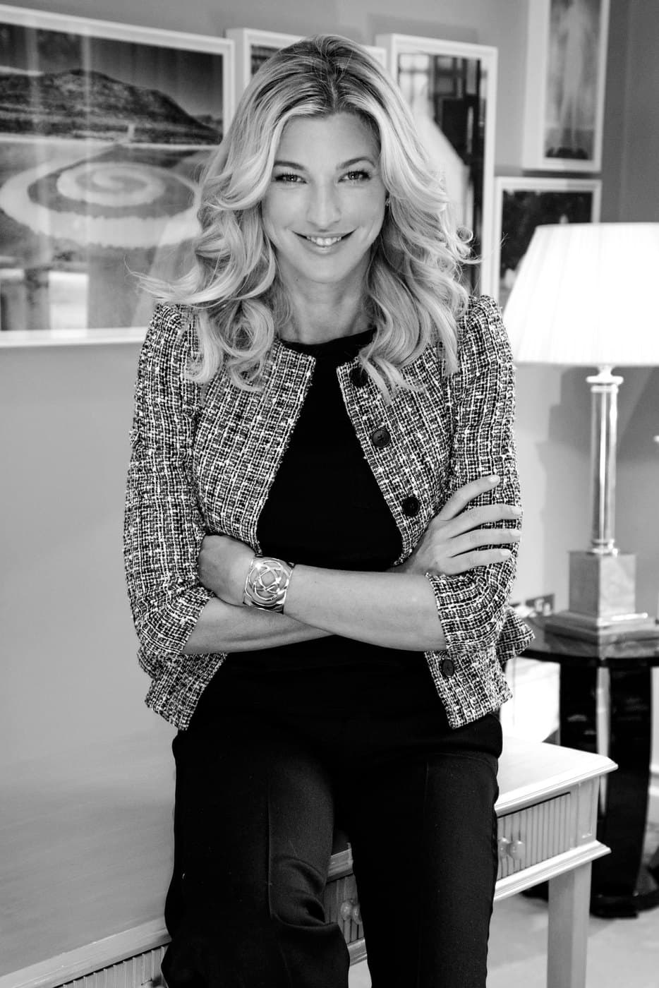 Nicole Junkerman in a tweed jacket with her arms folded, smiling in a bright room with framed pictures behind her, in black and white